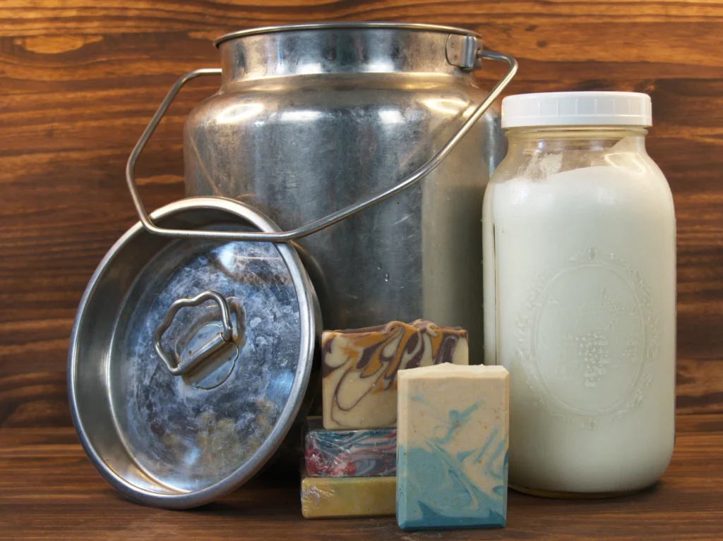 Fresh goat milk in a glass jar next to a stainless steel milk pail and handcrafted soap bars on a wooden background.